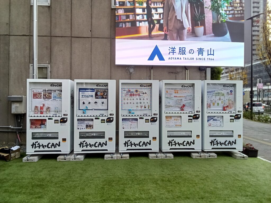 福山市伏見町の元福山東警察署駅前交番跡地「コウバンアトチ」に設置されたガチャCAN自動販売機の全景。5台が芝生敷きのスペースに並ぶ様子を正面北側から撮影したもので、駅前らしい開放的な雰囲気を示す。