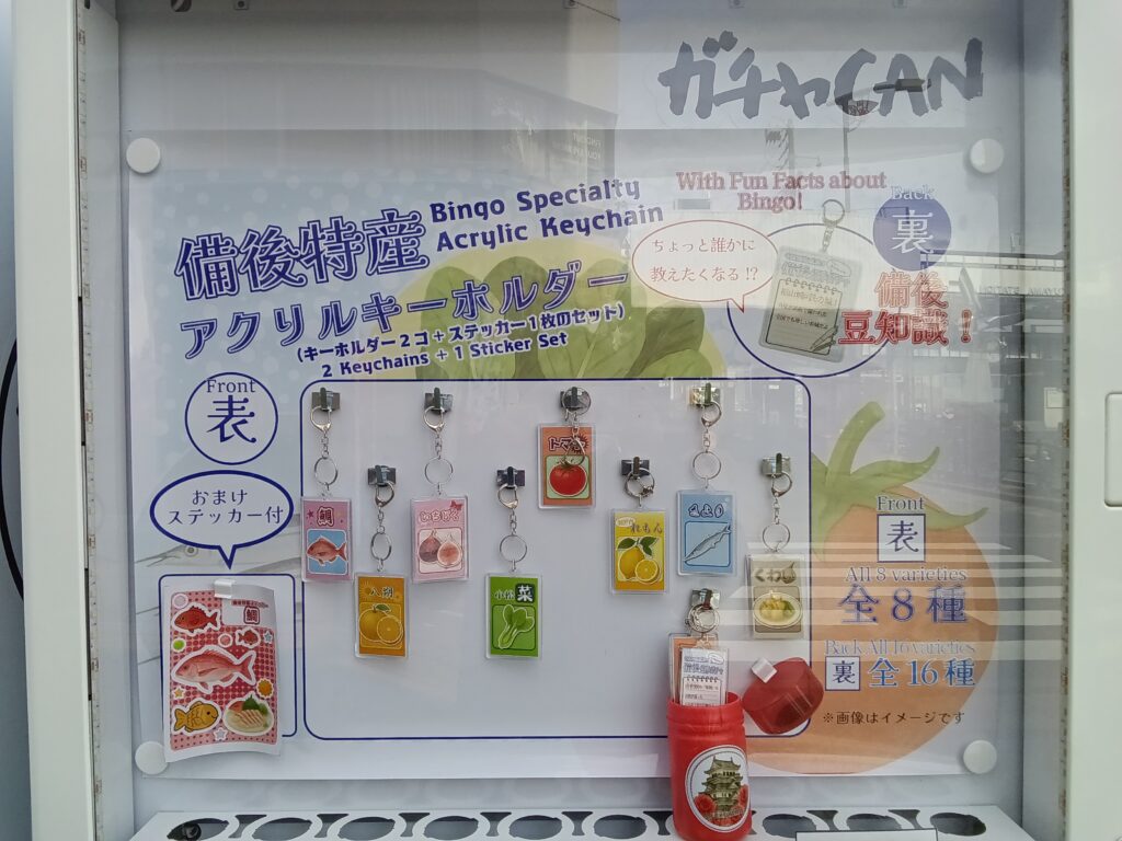 自販機内ディスプレイに並べられた福山周辺の特産品キーホルダー類。駅名標型アクリルや缶バッジなど、小売棚では出会いにくいラインナップの例を示す。